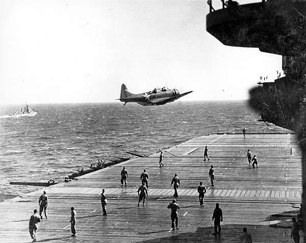 A scout plane passes low over Enterprise CV-6's flight deck to drop a weighted message, 18 May 1942.