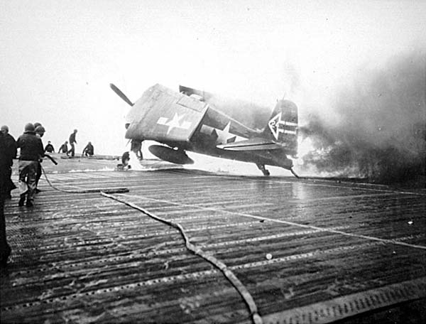 Damage control parties approach a VF(N)-90 Hellcat set afire by Kamikaze near-miss, 11 April 1945.